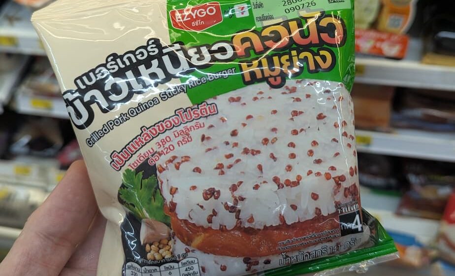 Ezygo Grilled Pork Quinoa Sticky Rice Burger: A Flavorful Street-Food Fusion with Serious Heat A hand holding a cream and green plastic pouch packaging of an Ezygo Grilled Pork Quinoa Sticky Rice Burger inside a store in Thailand.