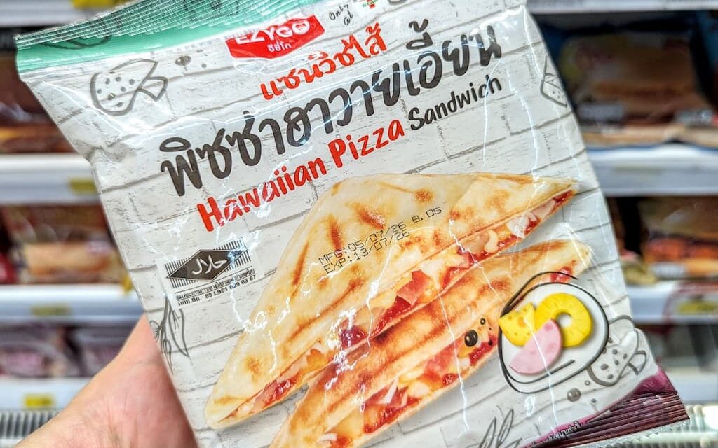 A hand holding a white plastic pouch packaging of an Ezygo Hawaiian Pizza Sandwich in front of store refrigerator shelves in Thailand.