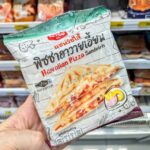 A hand holding a white plastic pouch packaging of an Ezygo Hawaiian Pizza Sandwich in front of store refrigerator shelves in Thailand.