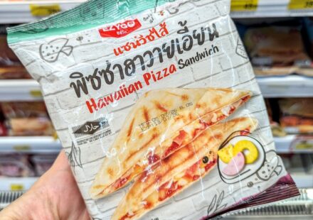 A hand holding a white plastic pouch packaging of an Ezygo Hawaiian Pizza Sandwich in front of store refrigerator shelves in Thailand.