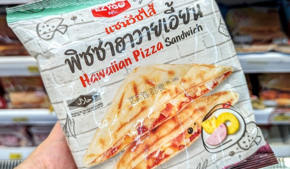 Ezygo Hawaiian Pizza Sandwich: A Crispy Toastie Weighed Down by Excessive Sweetness A hand holding a white plastic pouch packaging of an Ezygo Hawaiian Pizza Sandwich in front of store refrigerator shelves in Thailand.