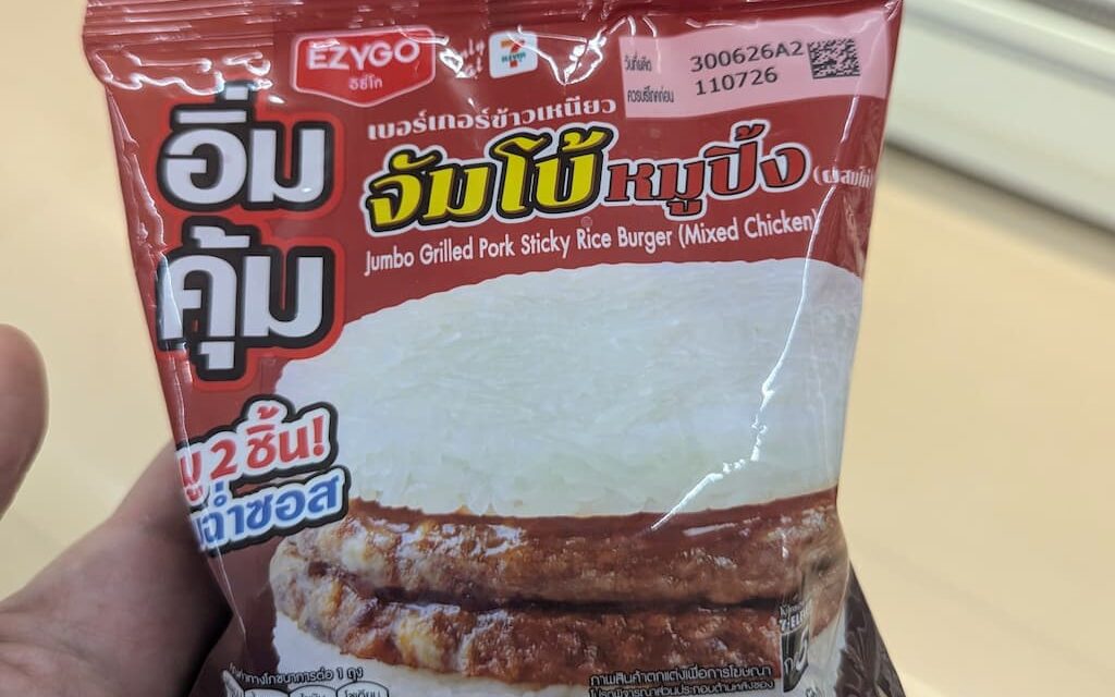 A hand holding a dark red plastic pouch packaging of an Ezygo Jumbo Grilled Pork Sticky Rice Burger inside a store in Thailand.