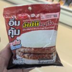 Are There 7-Elevens in Singapore? The Short Answer (And Why They Might Surprise You) A hand holding a dark red plastic pouch packaging of an Ezygo Jumbo Grilled Pork Sticky Rice Burger inside a store in Thailand.