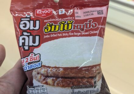 Ezygo Jumbo Grilled Pork Sticky Rice Burger: A Savory, High-Volume Street Fusion Done Right A hand holding a dark red plastic pouch packaging of an Ezygo Jumbo Grilled Pork Sticky Rice Burger inside a store in Thailand.