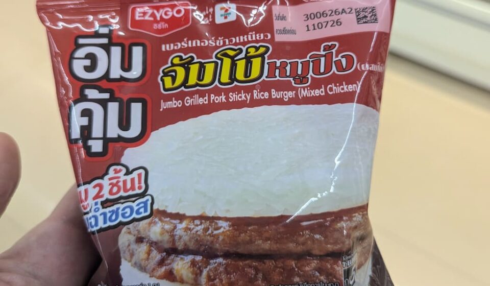 Ezygo Jumbo Grilled Pork Sticky Rice Burger: A Savory, High-Volume Street Fusion Done Right A hand holding a dark red plastic pouch packaging of an Ezygo Jumbo Grilled Pork Sticky Rice Burger inside a store in Thailand.