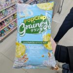 KitKat F1 Official Chocolate Bar: An Elite Racing-Themed Novelty That Delivers Outstanding Flavor A hand holding a light blue bag of Grainey Popchip Multigrain Sourcream & Onion inside a 7-Eleven store in Thailand.