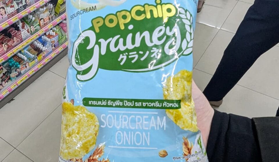 Grainey Popchip Multigrain Sourcream & Onion: A Light, Guilt-Free Snack That Scores on Subtle Flavor A hand holding a light blue bag of Grainey Popchip Multigrain Sourcream & Onion inside a 7-Eleven store in Thailand.