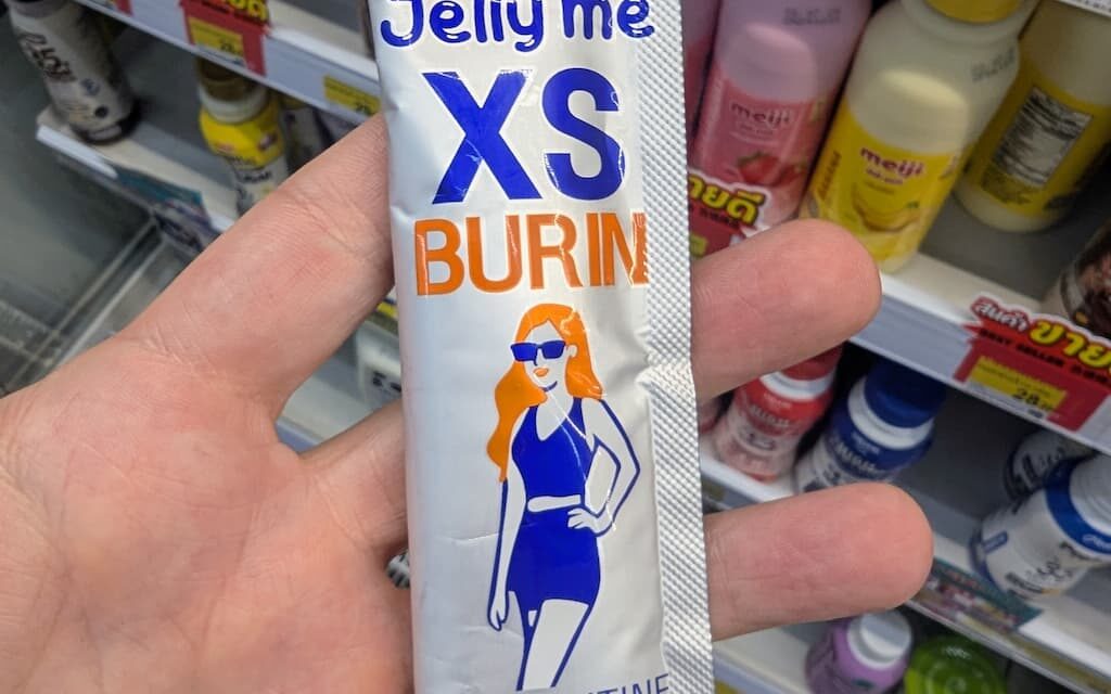 A hand holding a silver and orange single-serve stick pouch of HandyHerb Jelly Me XS Burin L-Carnitine on a store shelf in Thailand.