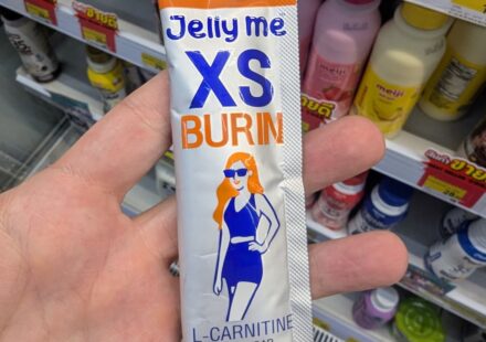 A hand holding a silver and orange single-serve stick pouch of HandyHerb Jelly Me XS Burin L-Carnitine on a store shelf in Thailand.