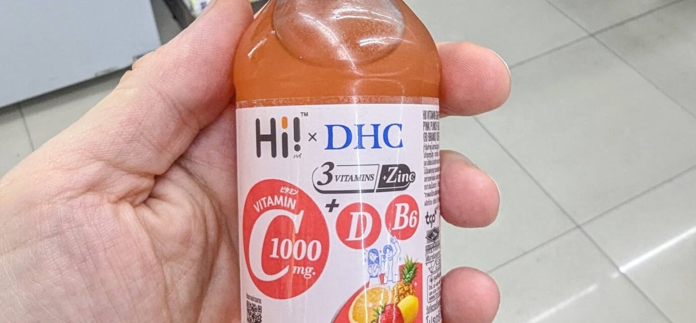 A hand holding a clear glass bottle of Hi! x DHC Vitamin C 1000mg Pink Punch drink with a royal blue cap inside a 7-Eleven in Thailand.