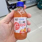A hand holding a clear glass bottle of Hi! x DHC Vitamin C 1000mg Pink Punch drink with a royal blue cap inside a 7-Eleven in Thailand.