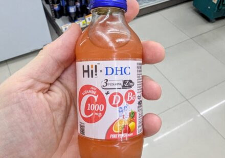 Hi! x DHC Vitamin C 1000mg Drink: The Elite Japanese-Formulated Immunity Shot A hand holding a clear glass bottle of Hi! x DHC Vitamin C 1000mg Pink Punch drink with a royal blue cap inside a 7-Eleven in Thailand.
