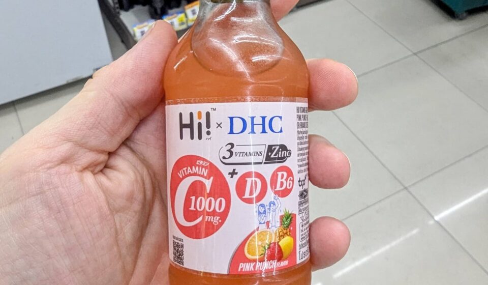 Hi! x DHC Vitamin C 1000mg Drink: The Elite Japanese-Formulated Immunity Shot A hand holding a clear glass bottle of Hi! x DHC Vitamin C 1000mg Pink Punch drink with a royal blue cap inside a 7-Eleven in Thailand.