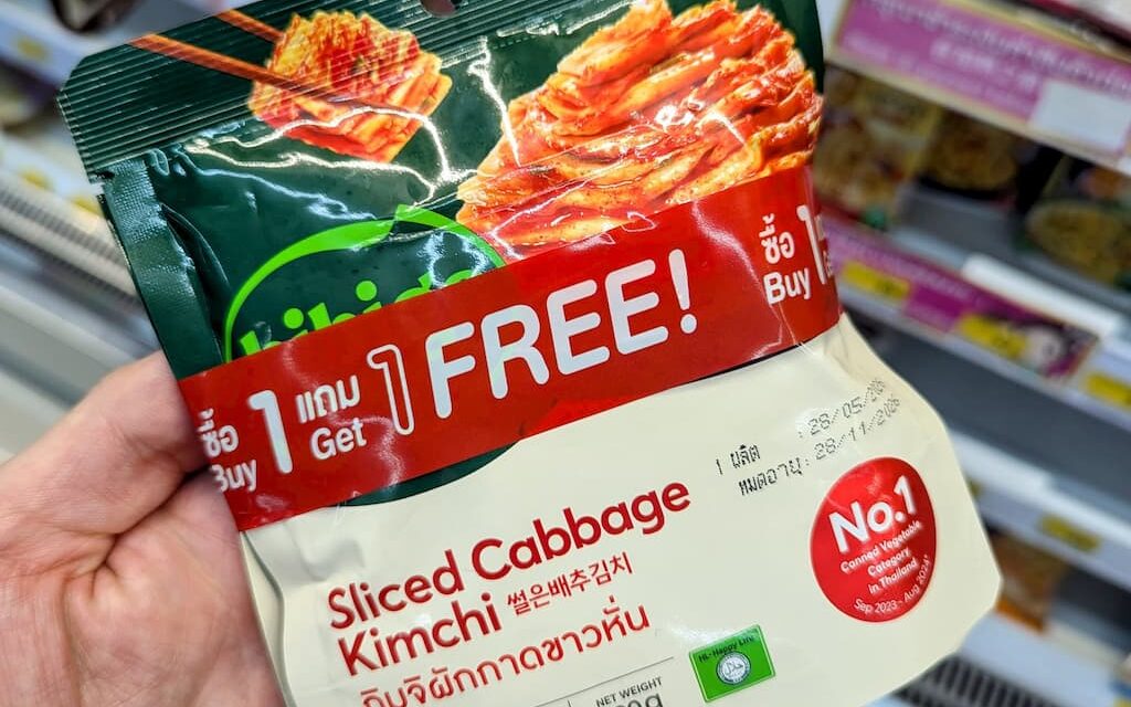 A hand holding a green and white pouch of King Chef Sliced Cabbage Kimchi with a red Buy 1 Get 1 Free banner inside a store in Thailand.