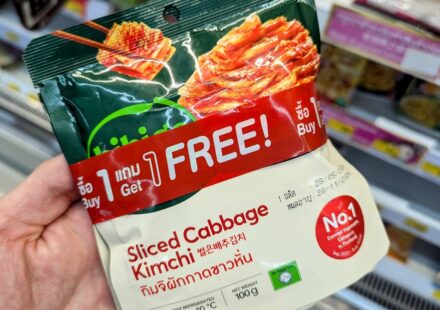 King Chef Sliced Cabbage Kimchi: The Essential Korean Side Dish for 7-Eleven Meals A hand holding a green and white pouch of King Chef Sliced Cabbage Kimchi with a red Buy 1 Get 1 Free banner inside a store in Thailand.