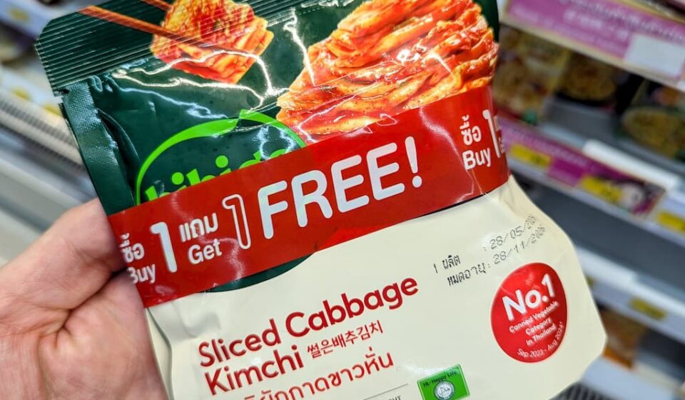 King Chef Sliced Cabbage Kimchi: The Essential Korean Side Dish for 7-Eleven Meals A hand holding a green and white pouch of King Chef Sliced Cabbage Kimchi with a red Buy 1 Get 1 Free banner inside a store in Thailand.