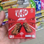 Hi! x DHC Vitamin C 1000mg Drink: The Elite Japanese-Formulated Immunity Shot A hand holding a red box of KitKat F1 Official Chocolate Bar featuring two molded chocolate racing cars inside a 7-Eleven in Thailand.