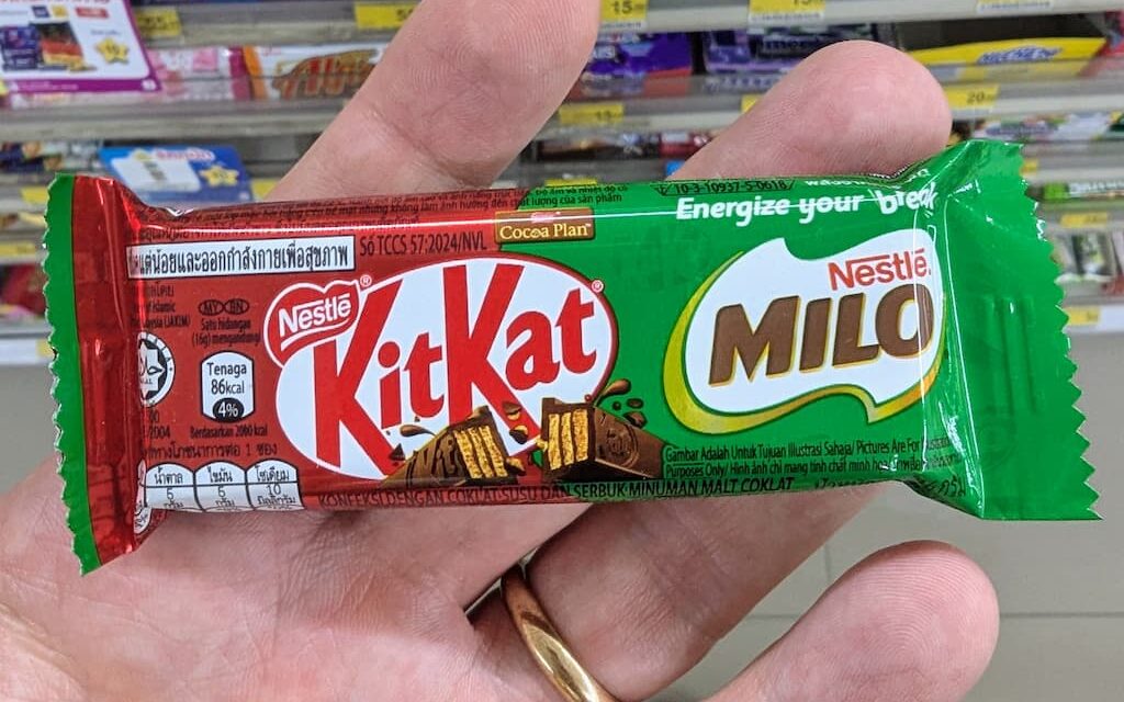 A hand holding a green KitKat x Milo single wafer chocolate bar inside a 7-Eleven store in Thailand.