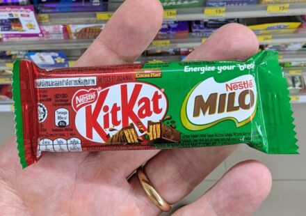 A hand holding a green KitKat x Milo single wafer chocolate bar inside a 7-Eleven store in Thailand.