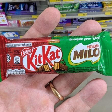 A hand holding a green KitKat x Milo single wafer chocolate bar inside a 7-Eleven store in Thailand.