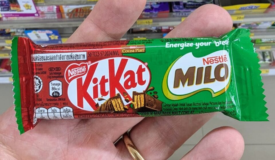 KitKat x Milo Chocolate Bar: A Malt-Infused Mashup Heavy on Sweetness A hand holding a green KitKat x Milo single wafer chocolate bar inside a 7-Eleven store in Thailand.