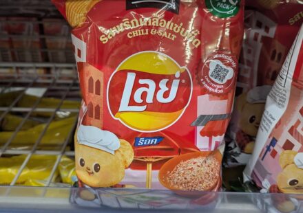 A hand holding a red packaging bag of Lay's Rock Chili and Salt Limited Edition potato chips on a 7-Eleven store shelf in Thailand.