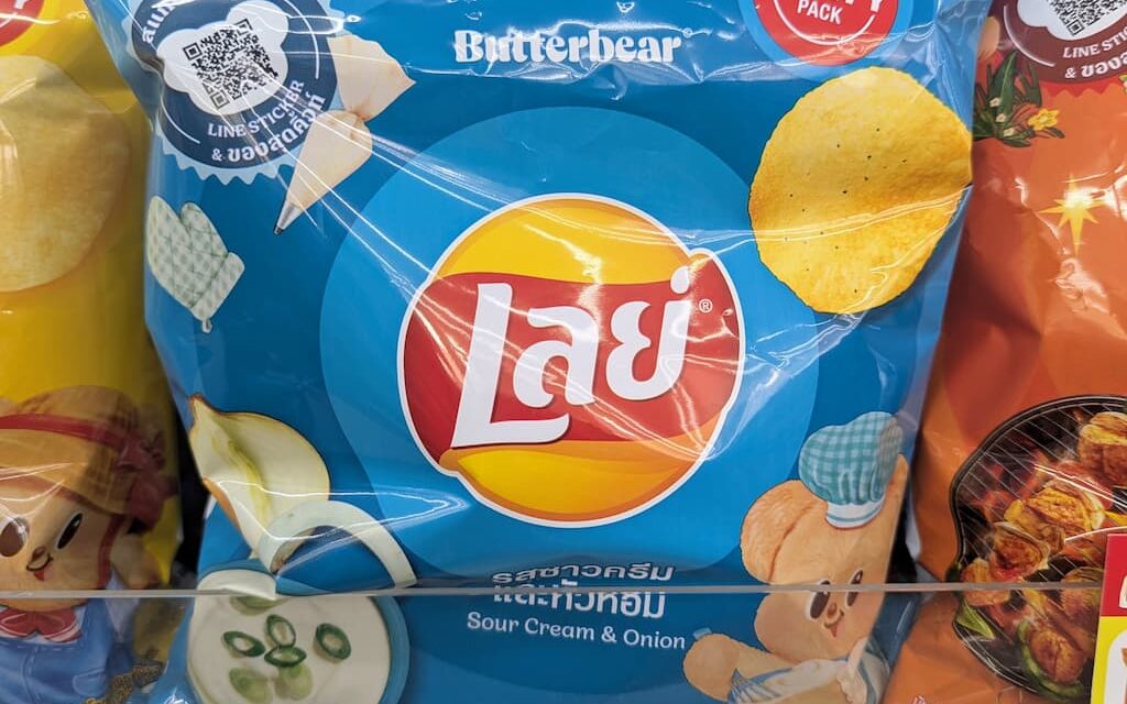 A hand holding a 105g blue bag of Lay's Sour Cream & Onion potato chips featuring Butterbear graphics inside a 7-Eleven store in Thailand.