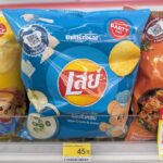 A hand holding a 105g blue bag of Lay's Sour Cream & Onion potato chips featuring Butterbear graphics inside a 7-Eleven store in Thailand.