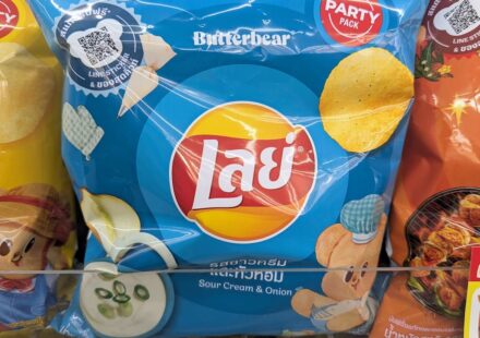 A hand holding a 105g blue bag of Lay's Sour Cream & Onion potato chips featuring Butterbear graphics inside a 7-Eleven store in Thailand.