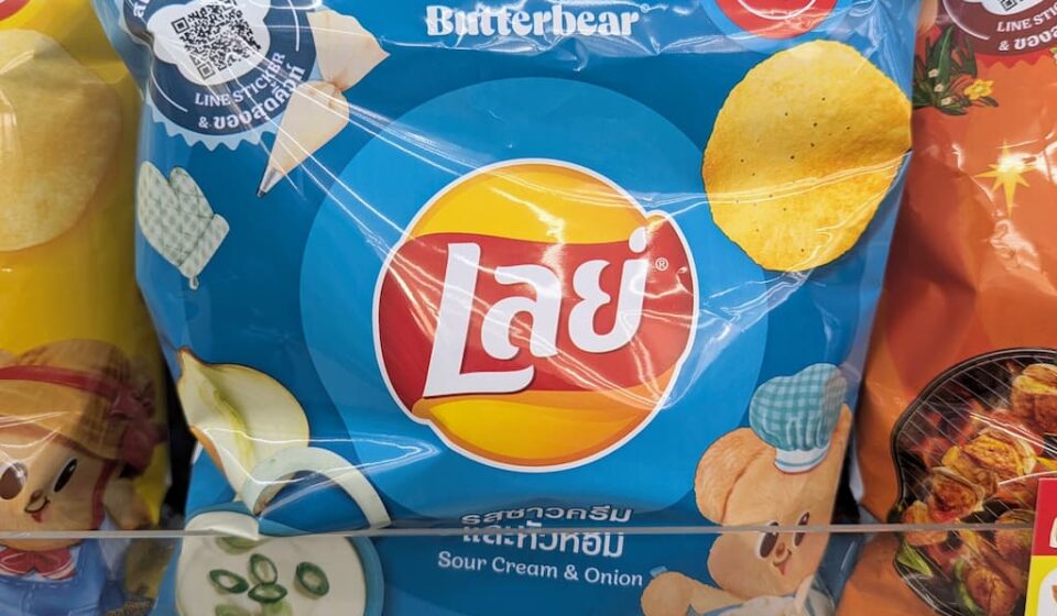 A hand holding a 105g blue bag of Lay's Sour Cream & Onion potato chips featuring Butterbear graphics inside a 7-Eleven store in Thailand.