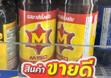 Two glass bottles of M-150 Original energy and vitamin drink behind a yellow 10-Baht price tag on a 7-Eleven store shelf in Thailand.