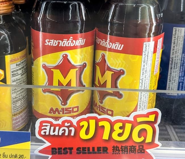 Two glass bottles of M-150 Original energy and vitamin drink behind a yellow 10-Baht price tag on a 7-Eleven store shelf in Thailand.