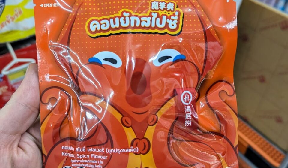 Magic Cook Konjac Spicy Flavour (Haidilao): A Fiery, Chewy Drinking Snack with Serious Heat A hand holding an orange pouch of Magic Cook Konjac Spicy Flavour by Haidilao featuring a cartoon octopus inside a 7-Eleven in Thailand.