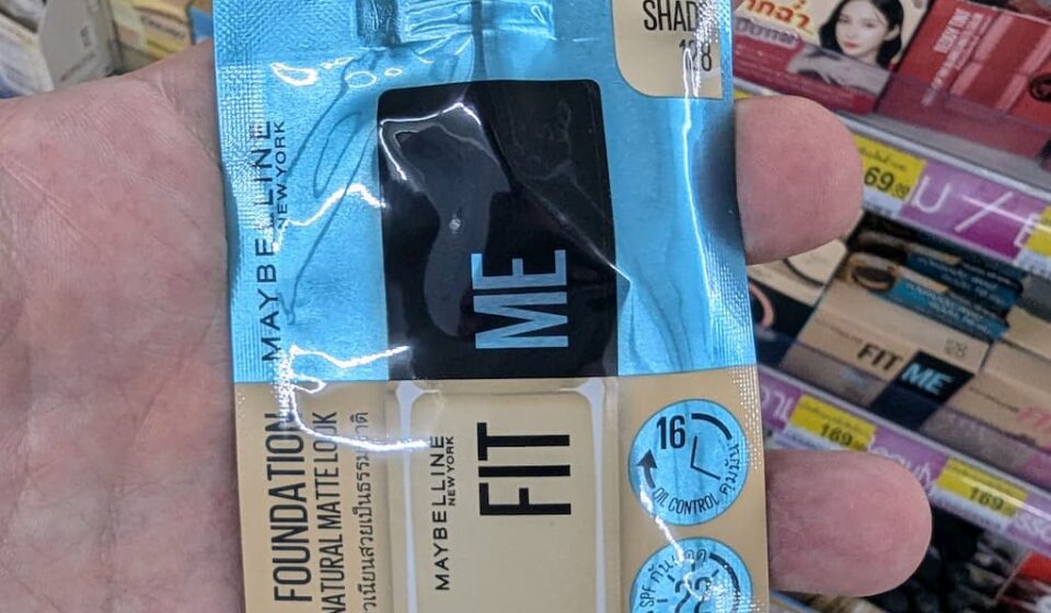 Maybelline Fit Me Matte + Poreless Foundation Sachet (Shade 128): Convenient Western Classic Facing Tough K-Beauty Competition A hand holding a Maybelline Fit Me Matte + Poreless Foundation sachet in Shade 128 inside a 7-Eleven store in Thailand.