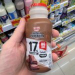 A hand holding a bottle of Meiji High Protein Chocolate Milk with 17g protein and no sugar added inside a 7-Eleven in Thailand.