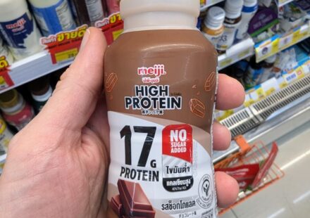 Meiji High Protein Chocolate Milk (17g Protein): A Rich, Low-Carb Satiating Fitness Drink A hand holding a bottle of Meiji High Protein Chocolate Milk with 17g protein and no sugar added inside a 7-Eleven in Thailand.