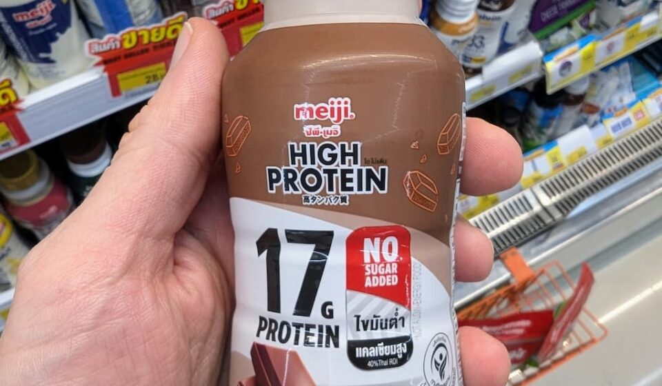 Meiji High Protein Chocolate Milk (17g Protein): A Rich, Low-Carb Satiating Fitness Drink A hand holding a bottle of Meiji High Protein Chocolate Milk with 17g protein and no sugar added inside a 7-Eleven in Thailand.