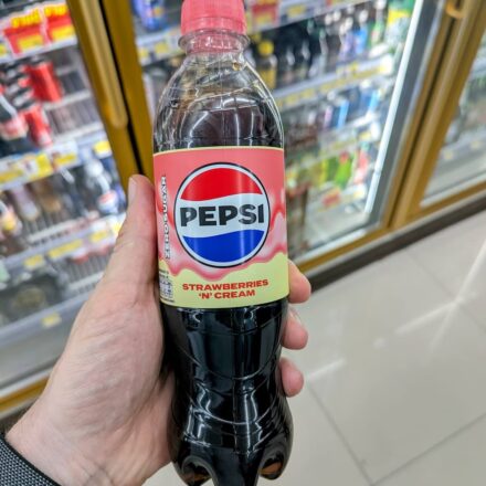 A hand holding a clear plastic bottle of Pepsi Strawberries 'N' Cream Zero Sugar soda with a pink cap inside a 7-Eleven store in Thailand.