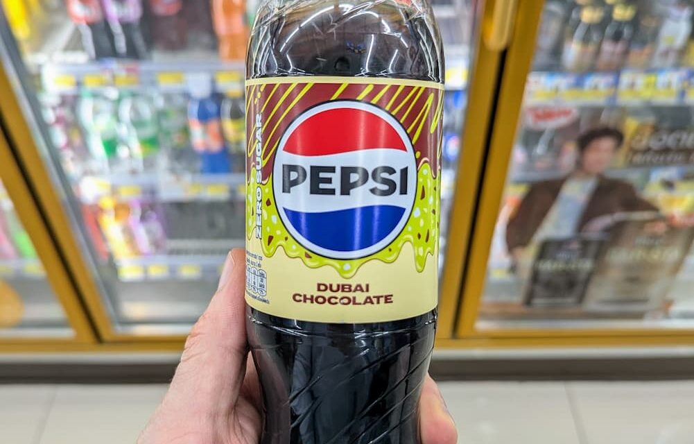 A hand holding a green-capped bottle of Pepsi Zero Sugar Dubai Chocolate flavor inside a 7-Eleven store in Thailand.