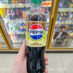 A hand holding a green-capped bottle of Pepsi Zero Sugar Dubai Chocolate flavor inside a 7-Eleven store in Thailand.