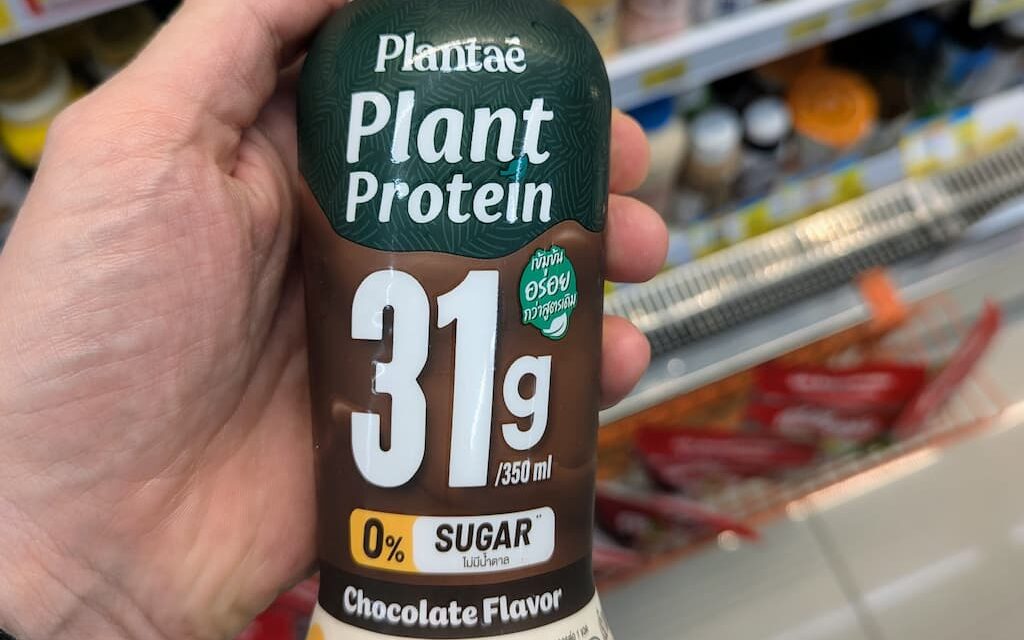A hand holding a bottle of Plantaē Plant Protein Chocolate Flavor with 31g protein inside a 7-Eleven in Thailand.