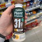 Who Owns 7-Eleven in Thailand? The Business Behind the Franchise A hand holding a bottle of Plantaē Plant Protein Chocolate Flavor with 31g protein inside a 7-Eleven in Thailand.