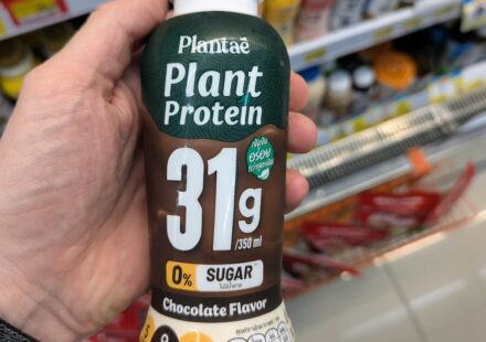 Plantaē Plant Protein Chocolate Flavor (31g Protein): Powerful Vegan Fuel with Rich Cocoa Taste A hand holding a bottle of Plantaē Plant Protein Chocolate Flavor with 31g protein inside a 7-Eleven in Thailand.