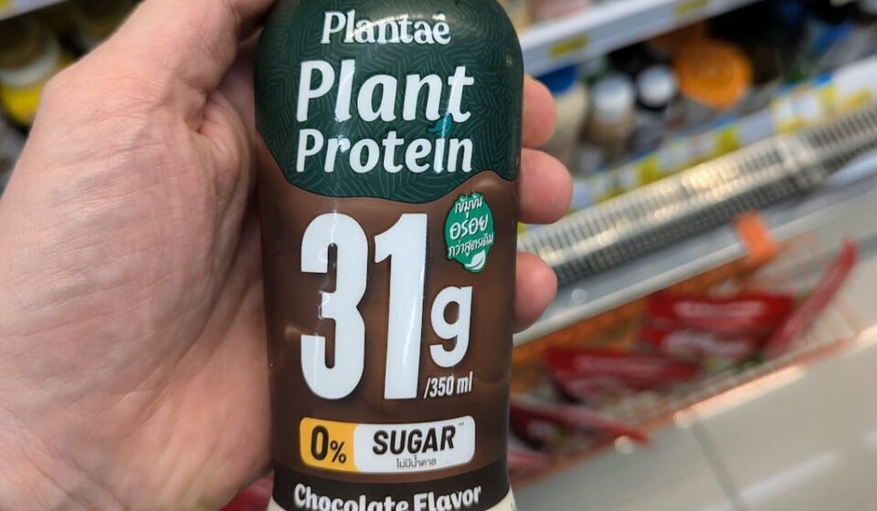 Plantaē Plant Protein Chocolate Flavor (31g Protein): Powerful Vegan Fuel with Rich Cocoa Taste A hand holding a bottle of Plantaē Plant Protein Chocolate Flavor with 31g protein inside a 7-Eleven in Thailand.