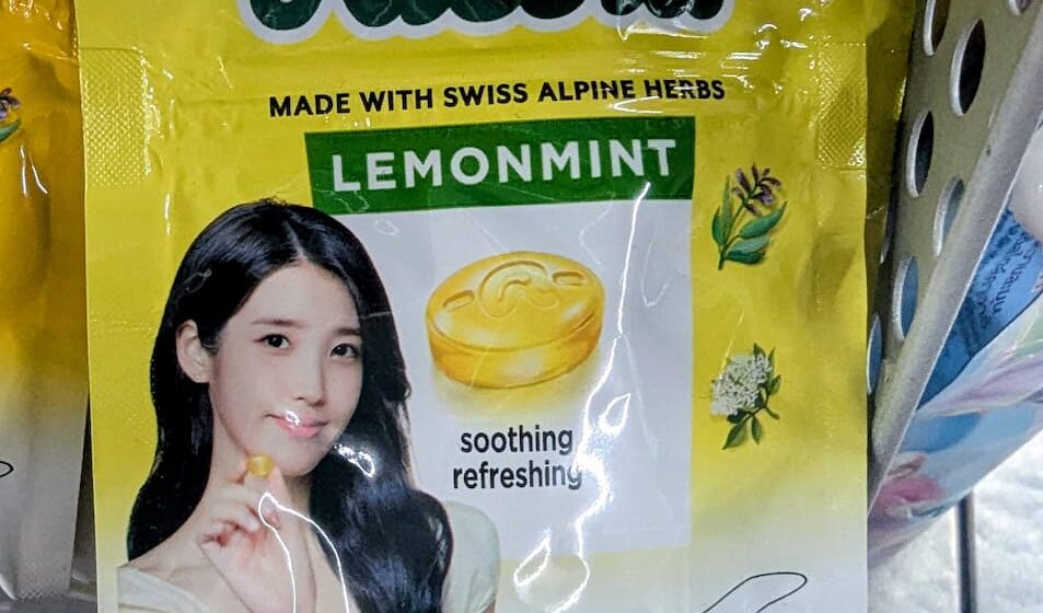 Ricola Lemonmint IU Edition: The Elite Swiss Herb Drop Powered by K-Pop Royalty A yellow hanging plastic pouch of limited edition Ricola Lemonmint sugar-free candy featuring K-pop idol IU on a store shelf in Thailand.