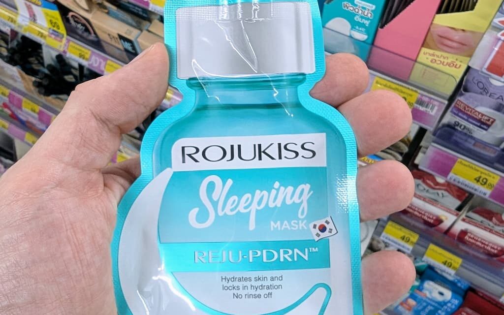 A hand holding a turquoise pouch of Rojukiss Reju-PDRN Sleeping Mask inside a 7-Eleven store in Thailand.