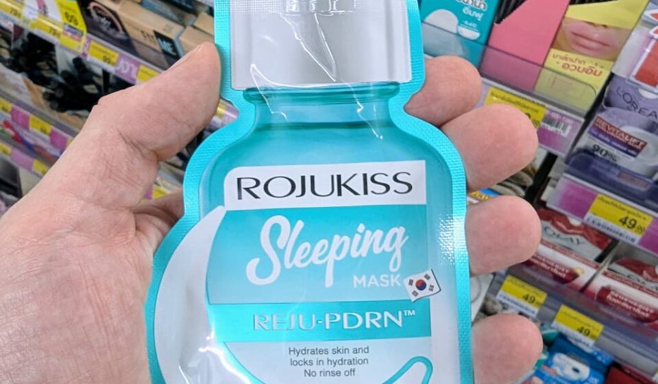 A hand holding a turquoise pouch of Rojukiss Reju-PDRN Sleeping Mask inside a 7-Eleven store in Thailand.