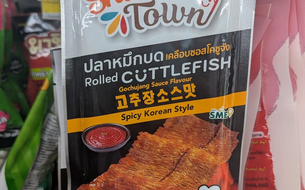 A hand holding a white and orange pouch of Snack Town Rolled Cuttlefish Gochujang Sauce Flavour inside a 7-Eleven in Thailand.