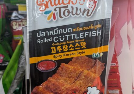 A hand holding a white and orange pouch of Snack Town Rolled Cuttlefish Gochujang Sauce Flavour inside a 7-Eleven in Thailand.