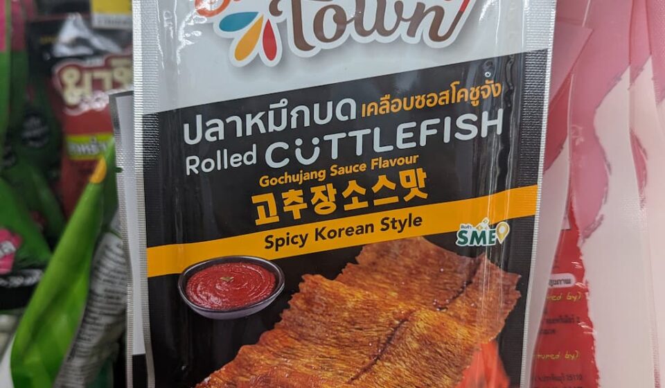 A hand holding a white and orange pouch of Snack Town Rolled Cuttlefish Gochujang Sauce Flavour inside a 7-Eleven in Thailand.